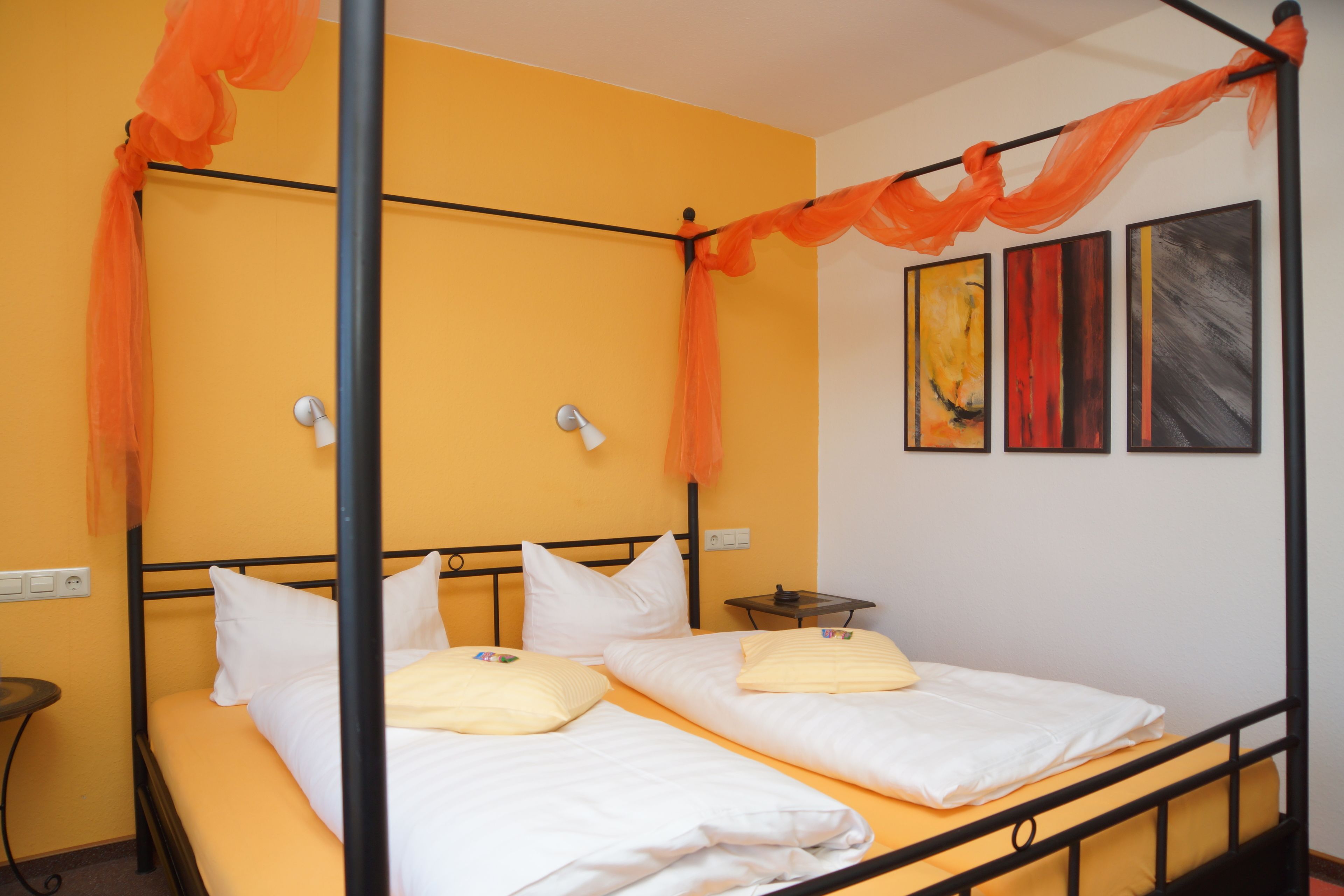 A room with a four-poster bed, two pillows, a bedside table, and a wall with three paintings.