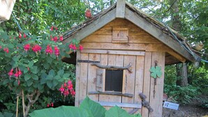 Une nichée à oiseaux en bois, dotée d’un toit incliné et d’une petite fenêtre, entourée d’un feuillage verdoyant et de fleurs rouge vif.