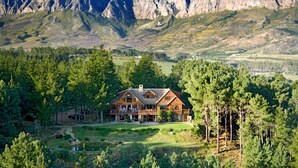 A large, two-story lodge with a prominent front porch and multiple windows, surrounded by dense forest and mountains in the background. - Lalapanzi Lodge (Cape Town)