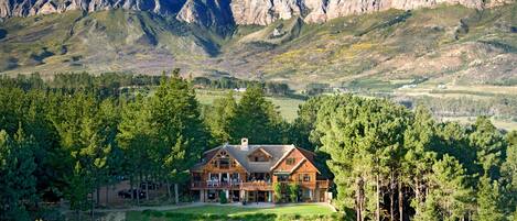A large, two-story lodge with a prominent front porch and multiple windows, surrounded by dense forest and mountains in the background.