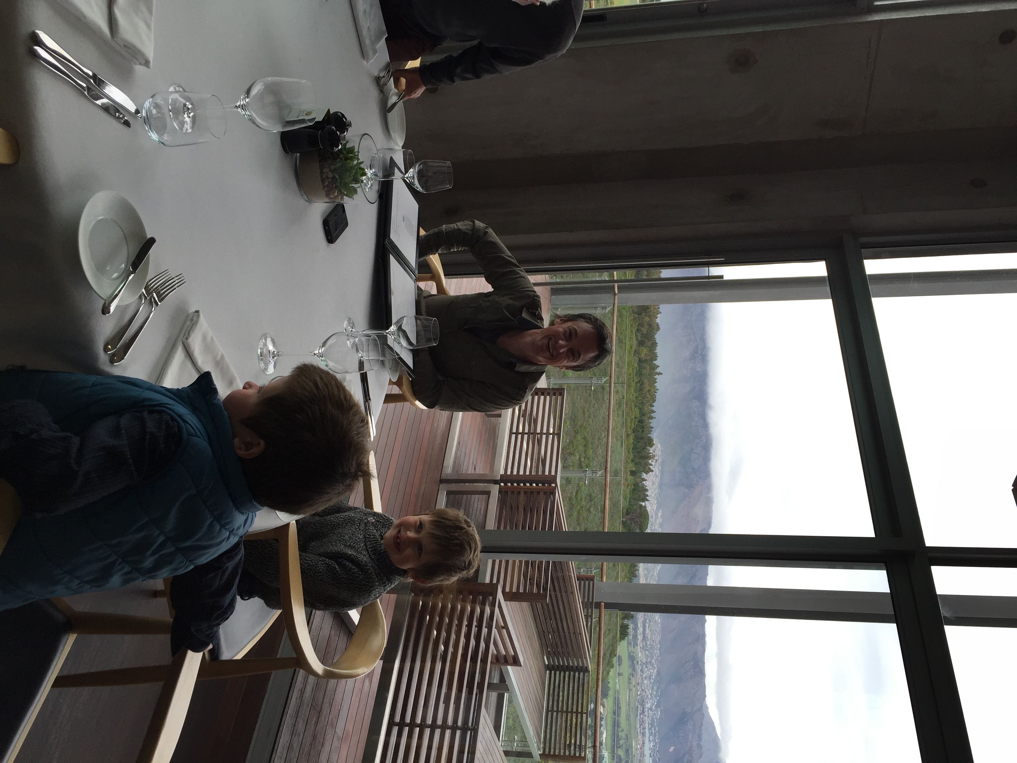 A dining table set with cutlery and wine glasses, with a view of a mountainous landscape through large windows.