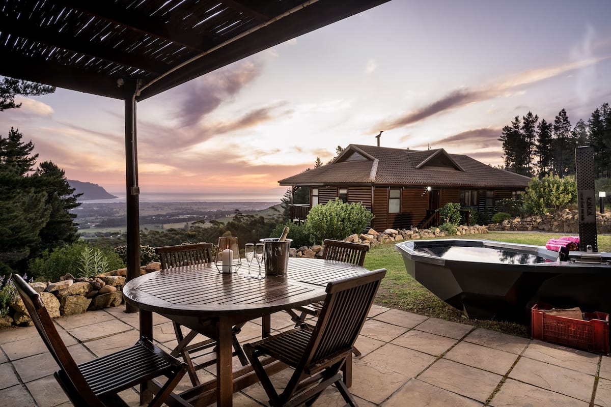 A wooden deck with a table and chairs, a hot tub, and a view of a mountain and sunset.