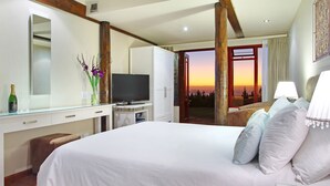 A hotel room with a bed, a desk with a TV, a mirror, and a view of a sunset through an open door. - Lalapanzi Lodge (Cape Town)