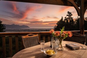 A wooden table with a glass of water, a bowl of food, a book, and a vase with flowers on a balcony overlooking a scenic view of mountains and a city at sunset. - Lalapanzi Lodge (Cape Town)