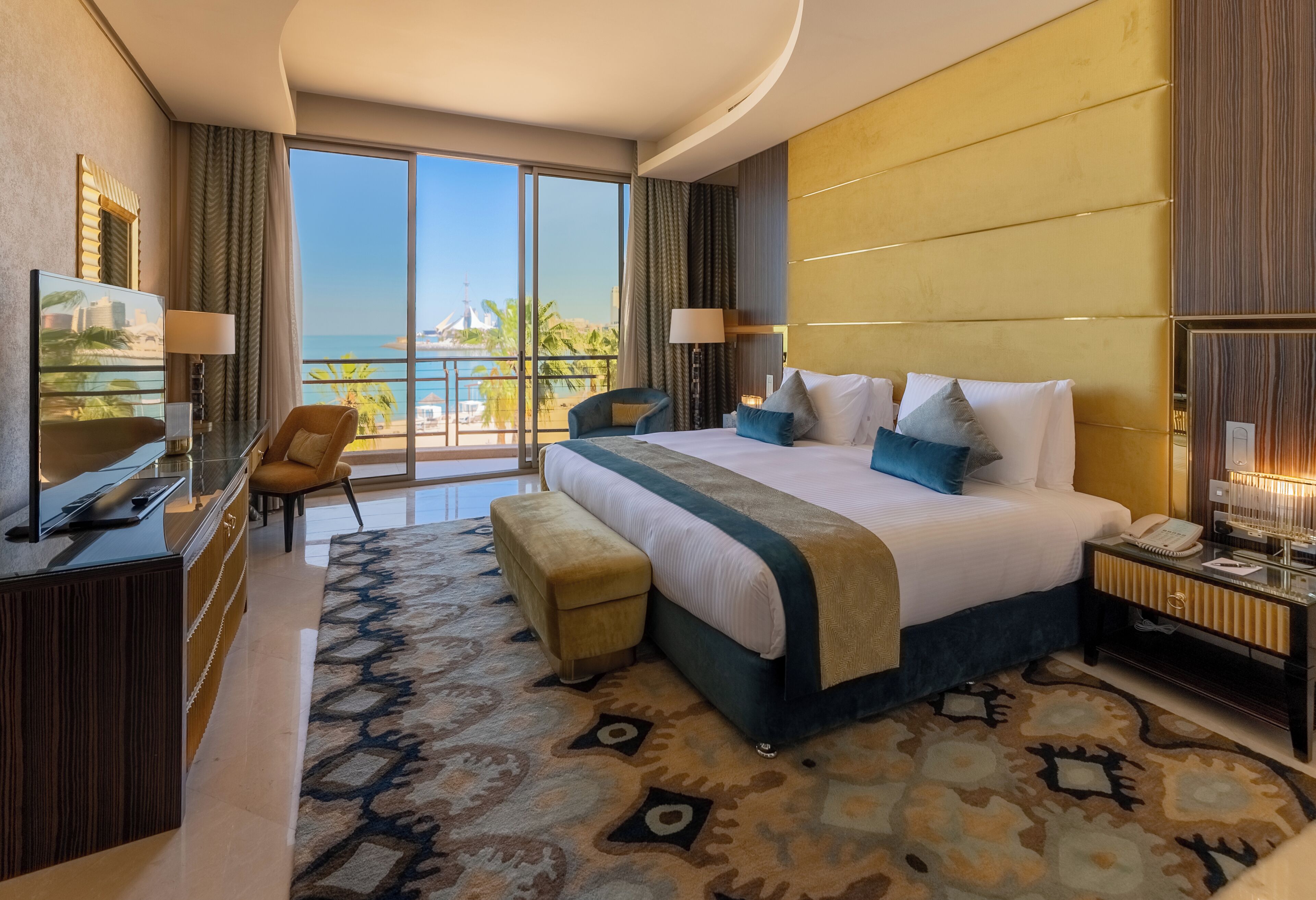 A hotel room with a large bed, a desk, a TV, and a balcony with a view of the sea.
