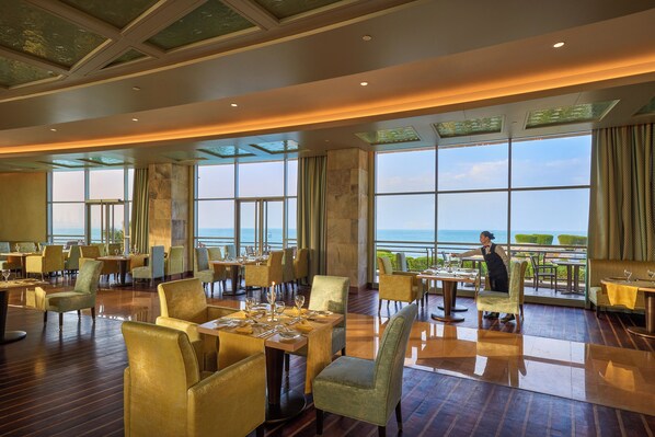 Un restaurante espacioso con mesas listas para cenar, grandes ventanales que ofrecen vista al mar y un techo alto con tragaluces.