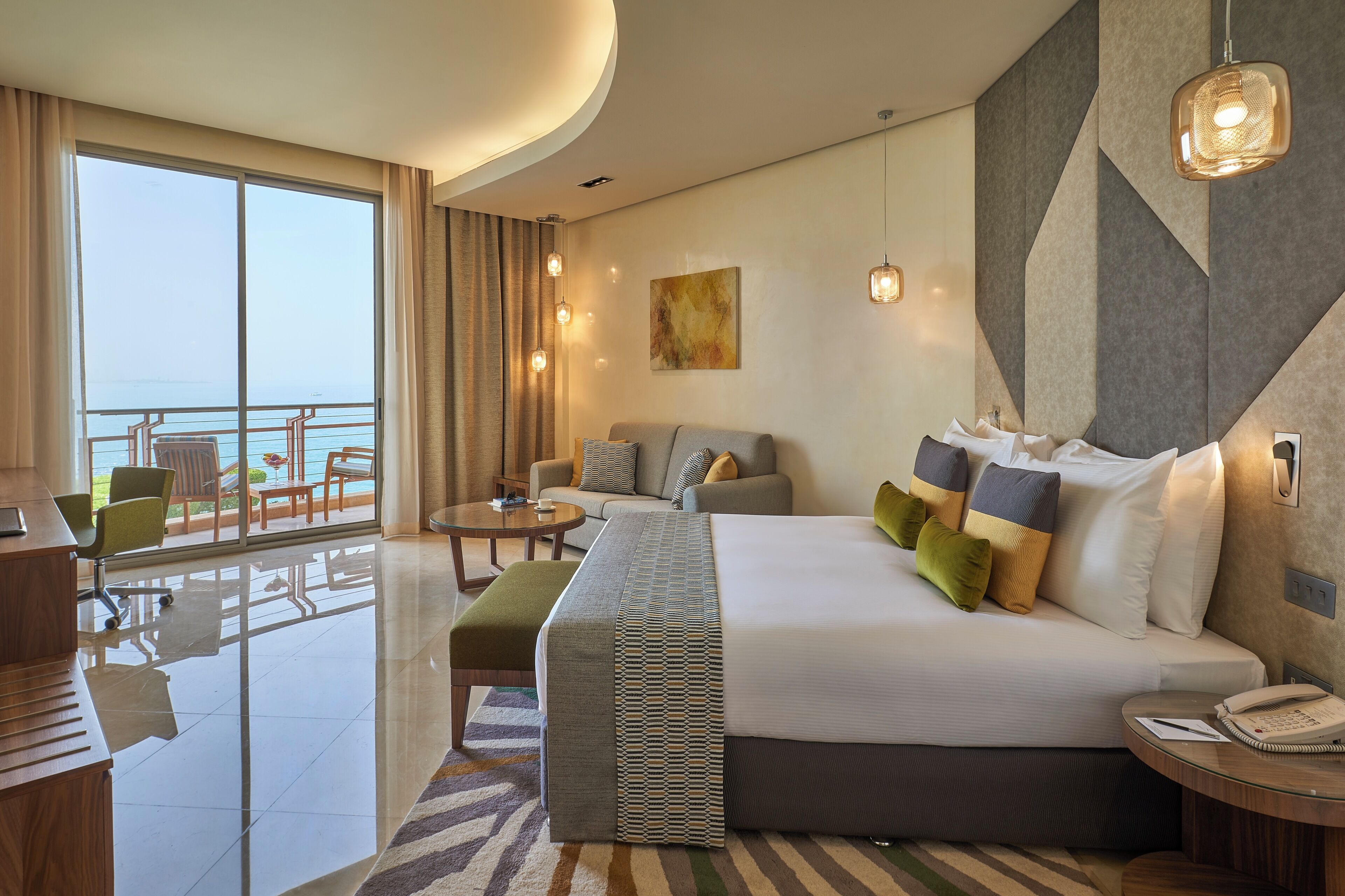 A hotel room with a large bed, a desk, a chair, a sofa, a balcony with a railing, and a view of the sea.
