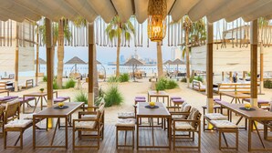 En restaurant ved stranden med træborde og stole, hængende lys og udsigt til havet og byens skyline.