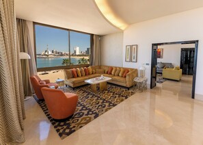 A spacious living room with a large window offering a view of a city skyline and water. There is a beige sofa, a red armchair, and a coffee table. - Marina Hotel Kuwait (Salmiya)