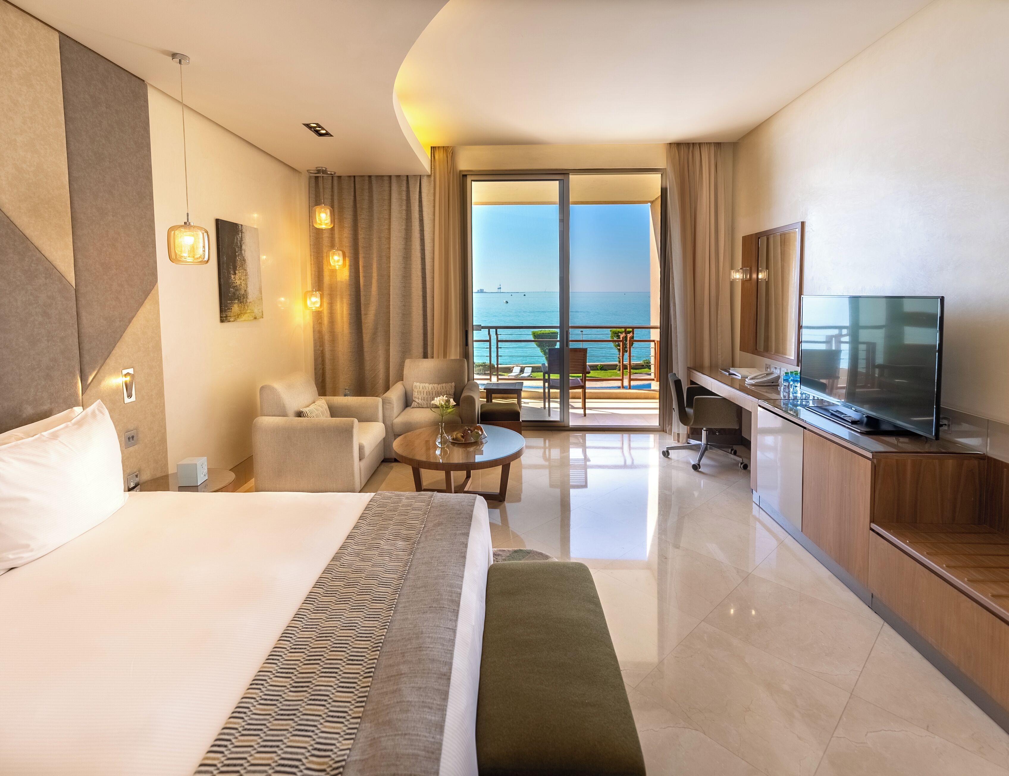 A hotel room with a large bed, a desk, a TV, and a balcony with a view of the sea.