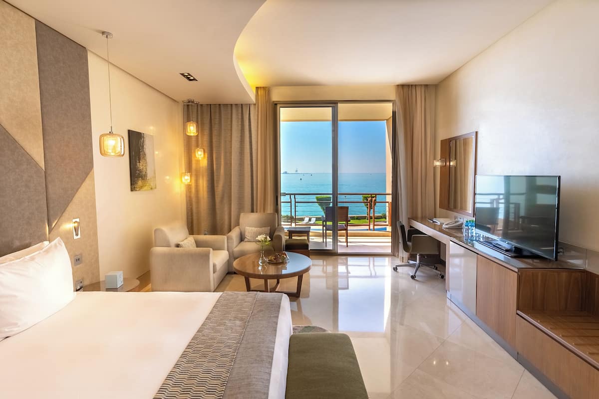 A hotel room with a large bed, a desk, a TV, and a balcony with a view of the sea.