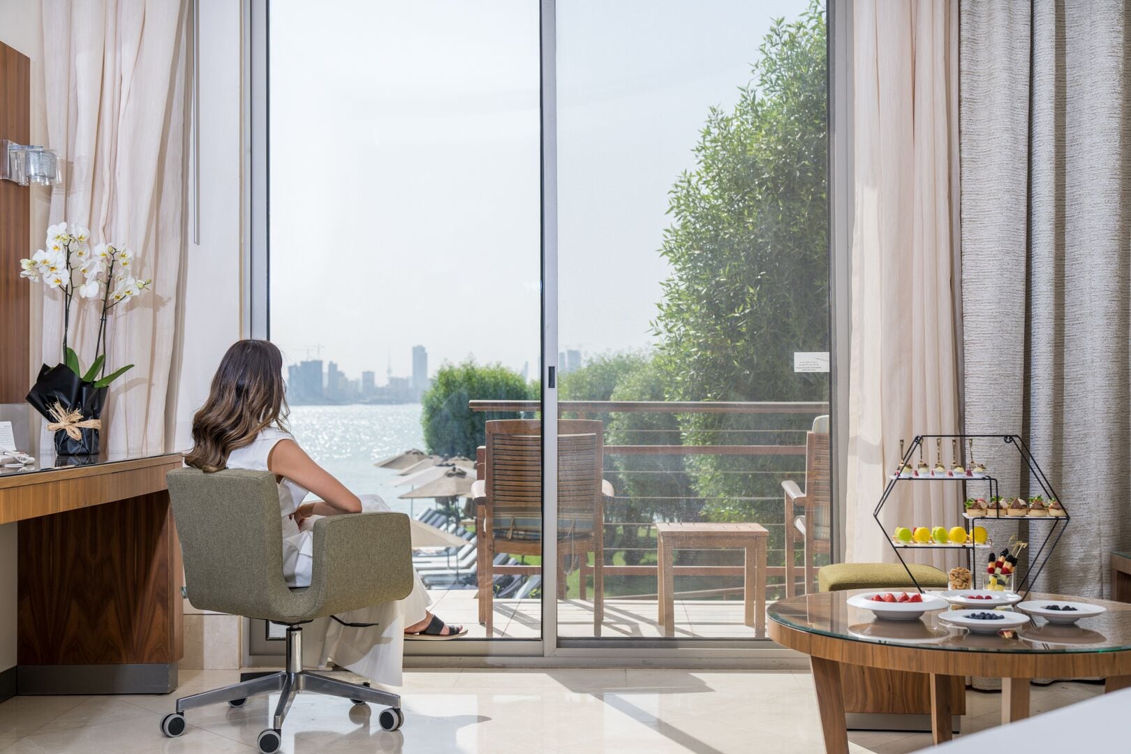 A modern room with a large window overlooking a cityscape and water. There is a desk with a computer, a chair, and a small table with fruit.