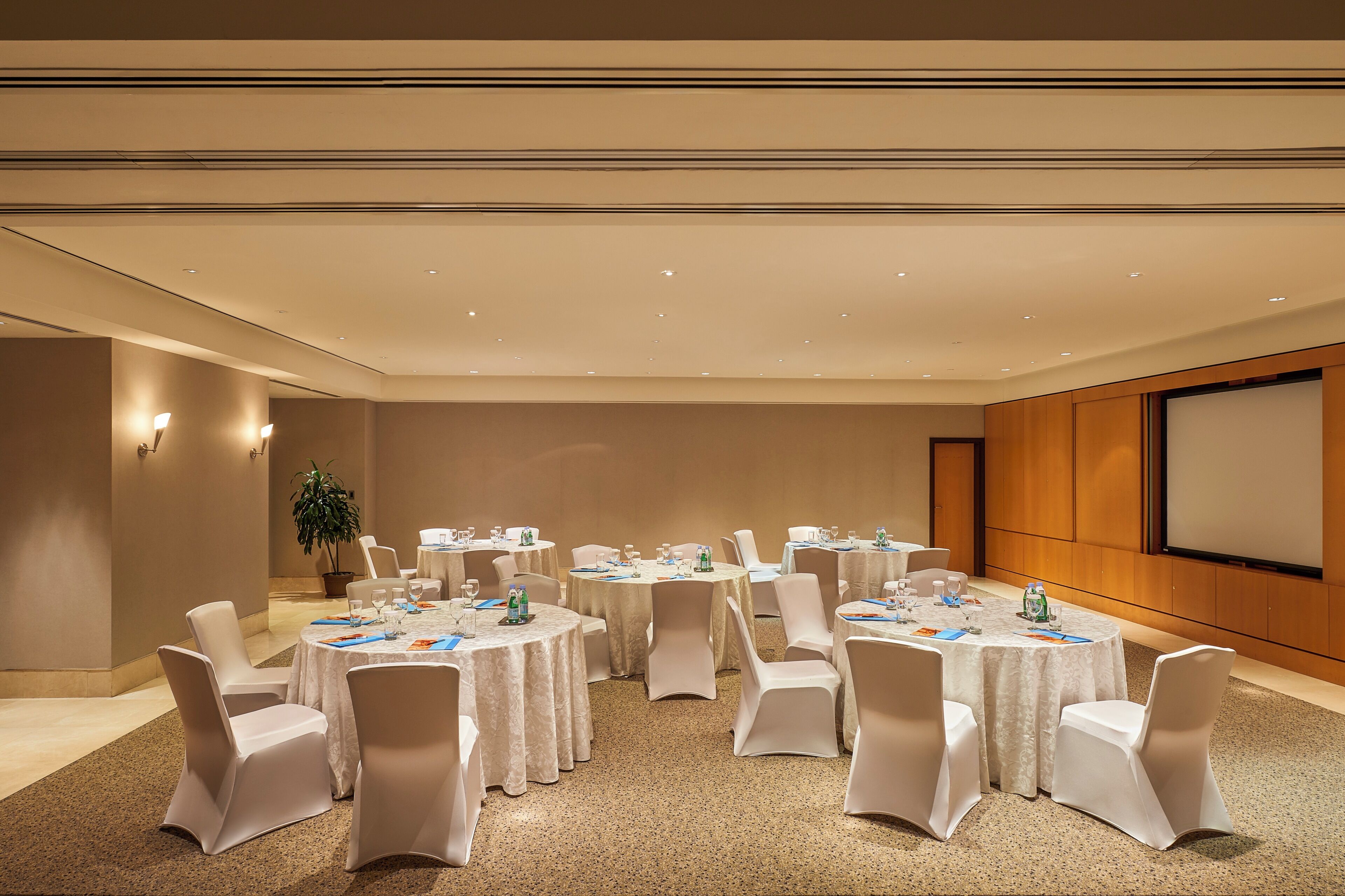 Un salón de conferencias con mesas redondas, sillas blancas y una pantalla de proyector.