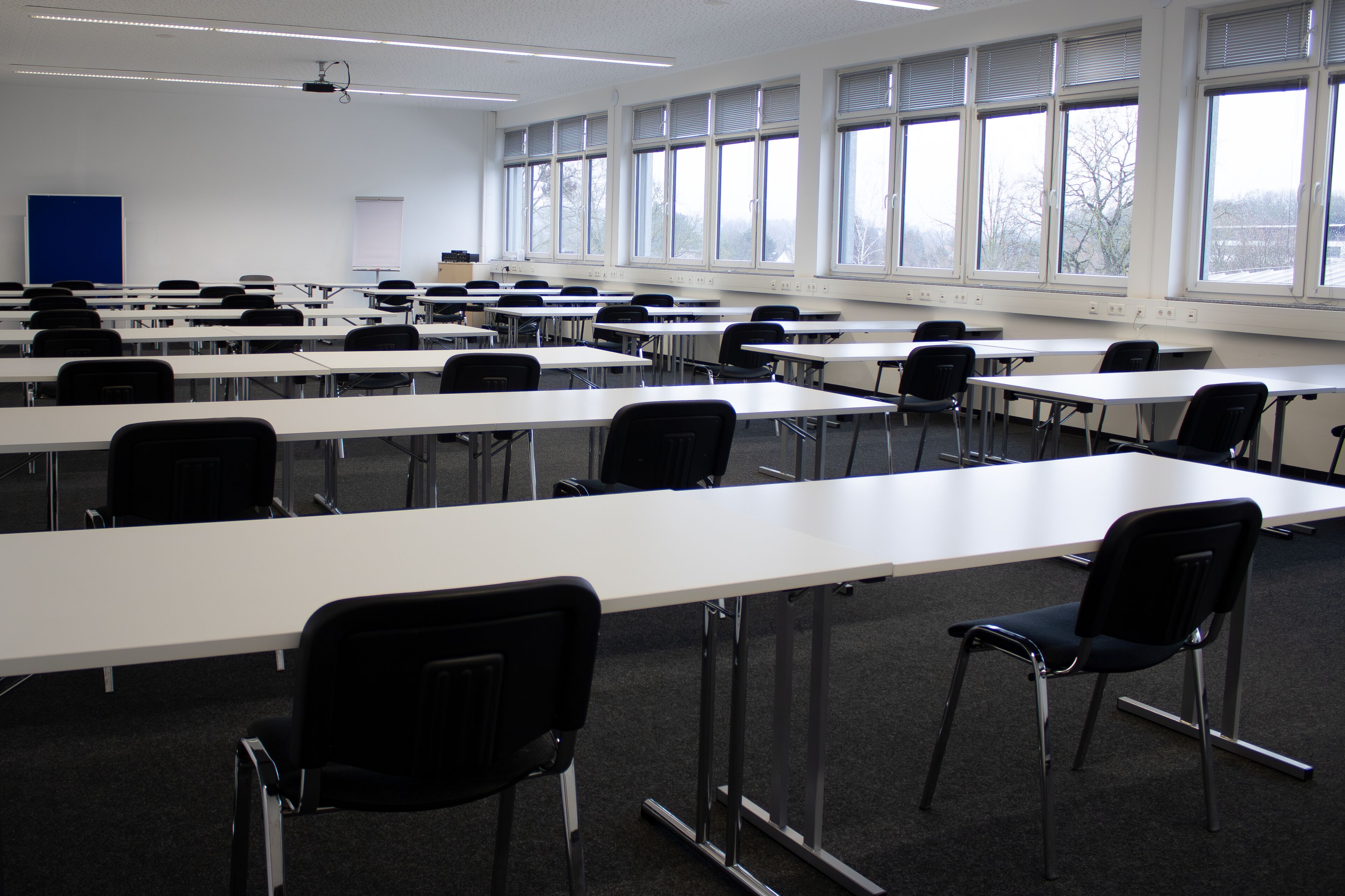 A classroom with rows of tables and chairs, large windows, and a projector on the ceiling.