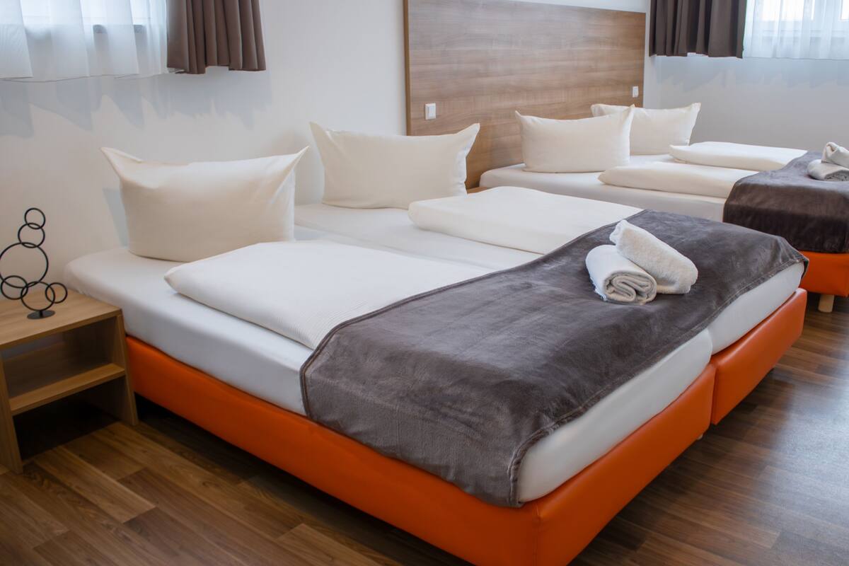 A hotel room with a large bed, a smaller bed, and a sofa bed. There is a wooden headboard, a wooden floor, and a window with curtains.