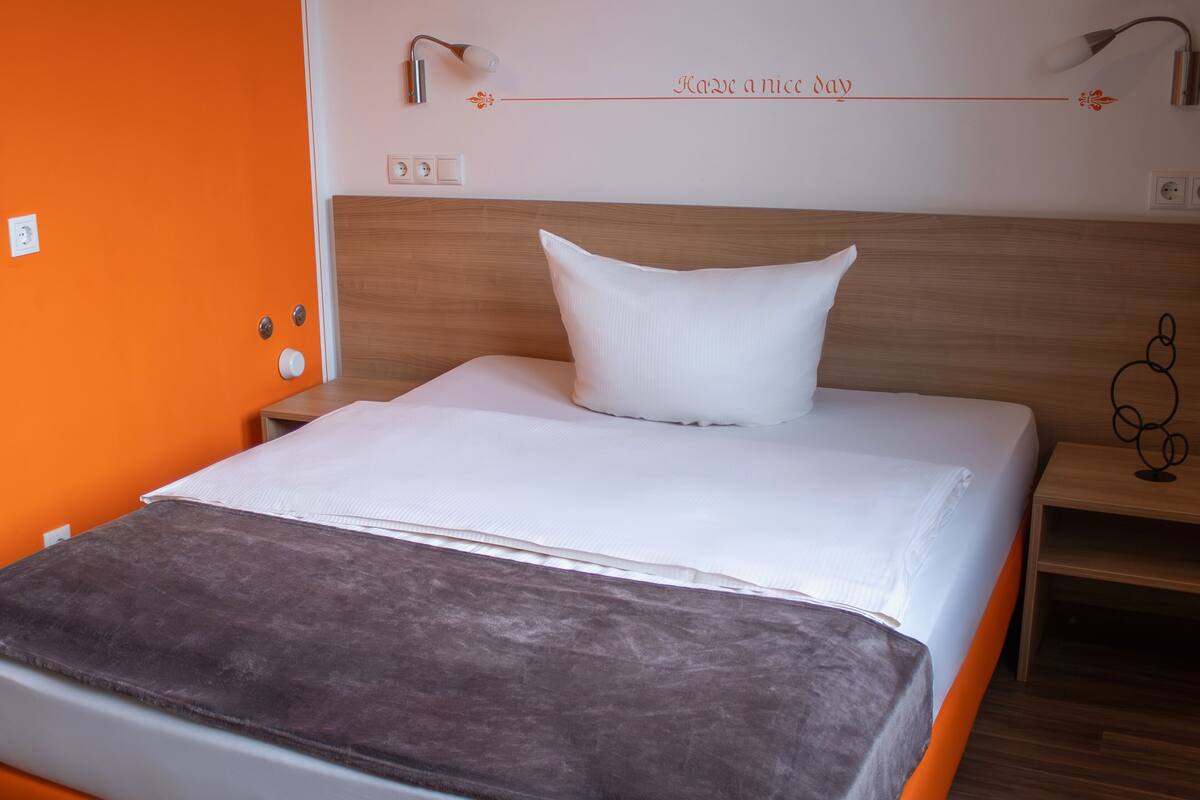 A neatly made bed with a white pillow and a brown blanket in a room with orange walls and a wooden headboard.