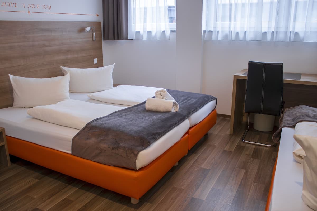 A hotel room with a wooden headboard, a bed with white and orange bedding, a desk with a chair, and a window with sheer curtains.