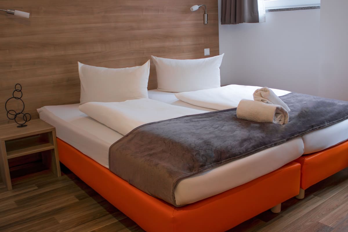 A modern hotel room with a wooden headboard, a bed with white linens and a grey blanket, an orange accent wall, and a small bedside table with a lamp.