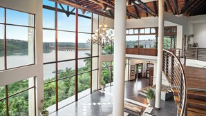 A spacious interior with large windows offering a view of a river and bridge, a modern staircase, and a cozy seating area with a fireplace. - Jozini Tiger Lodge by Dream Resorts (Jozini)