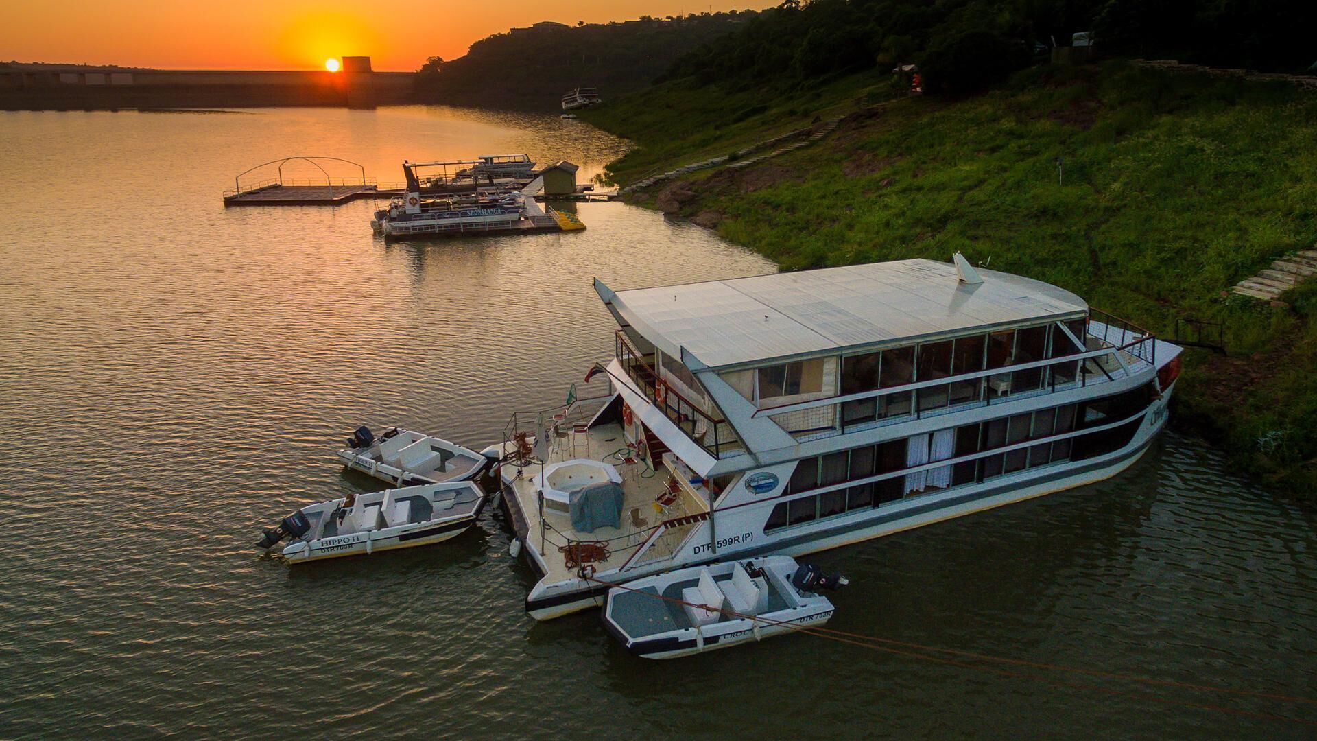 A large houseboat docked at a marina with multiple smaller boats nearby, surrounded by greenery and a sunset in the background.