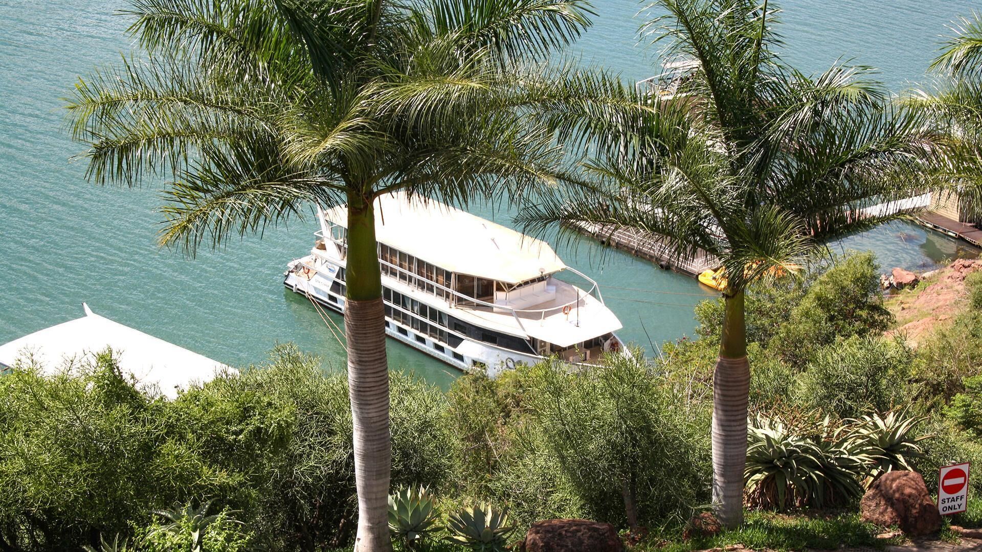 Une maison de plaisance avec un balcon, entourée de palmiers et donnant sur une étendue d’eau.
