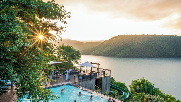 A swimming pool with people, surrounded by lush greenery and overlooking a lake with mountains in the background. - Jozini Tiger Lodge by Dream Resorts (Jozini)
