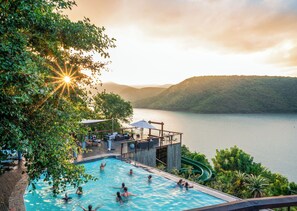 A swimming pool with people, surrounded by lush greenery and overlooking a lake with mountains in the background.