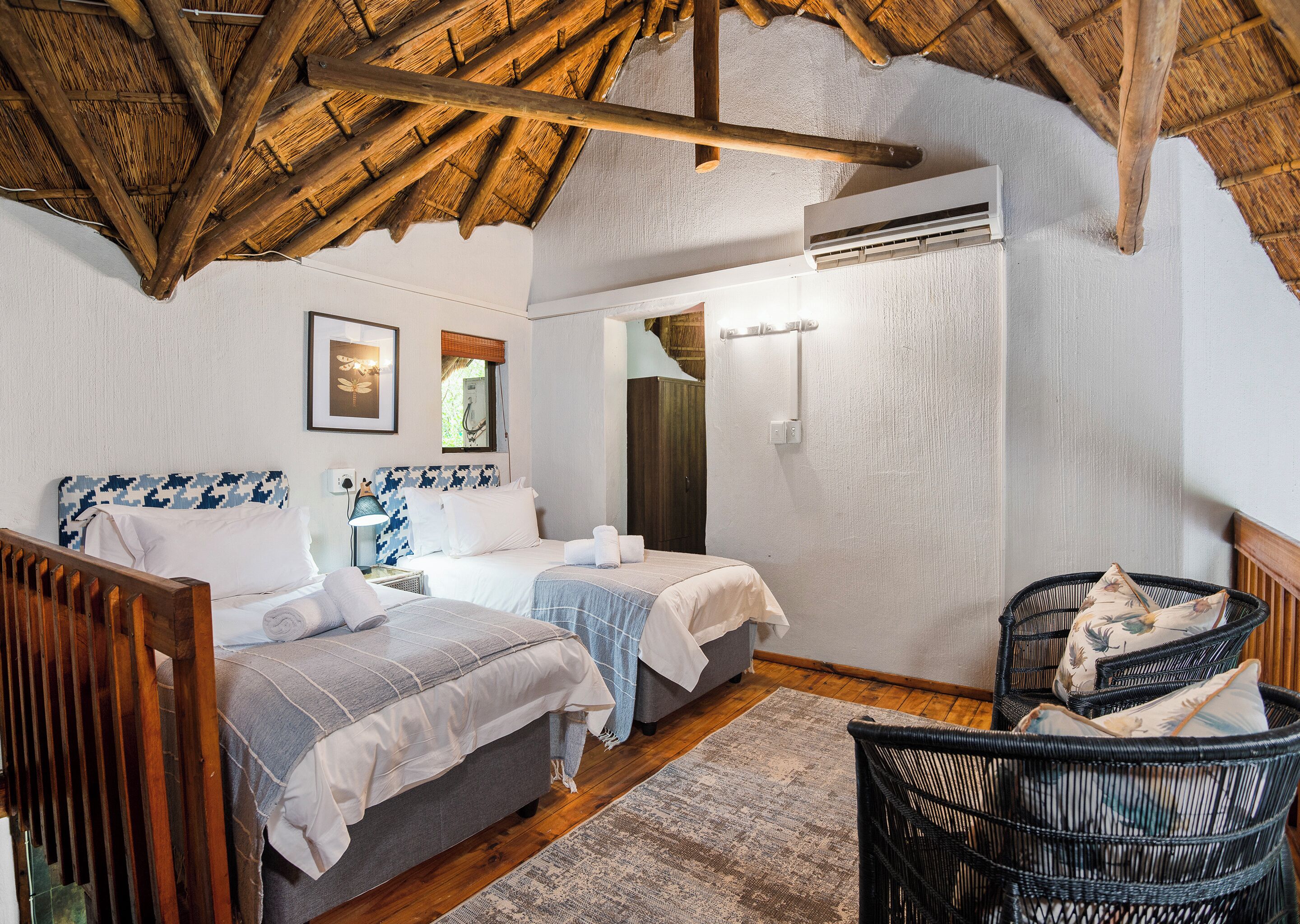 A room with two beds, a wooden ceiling, an air conditioning unit, a wicker chair, and a framed picture on the wall.