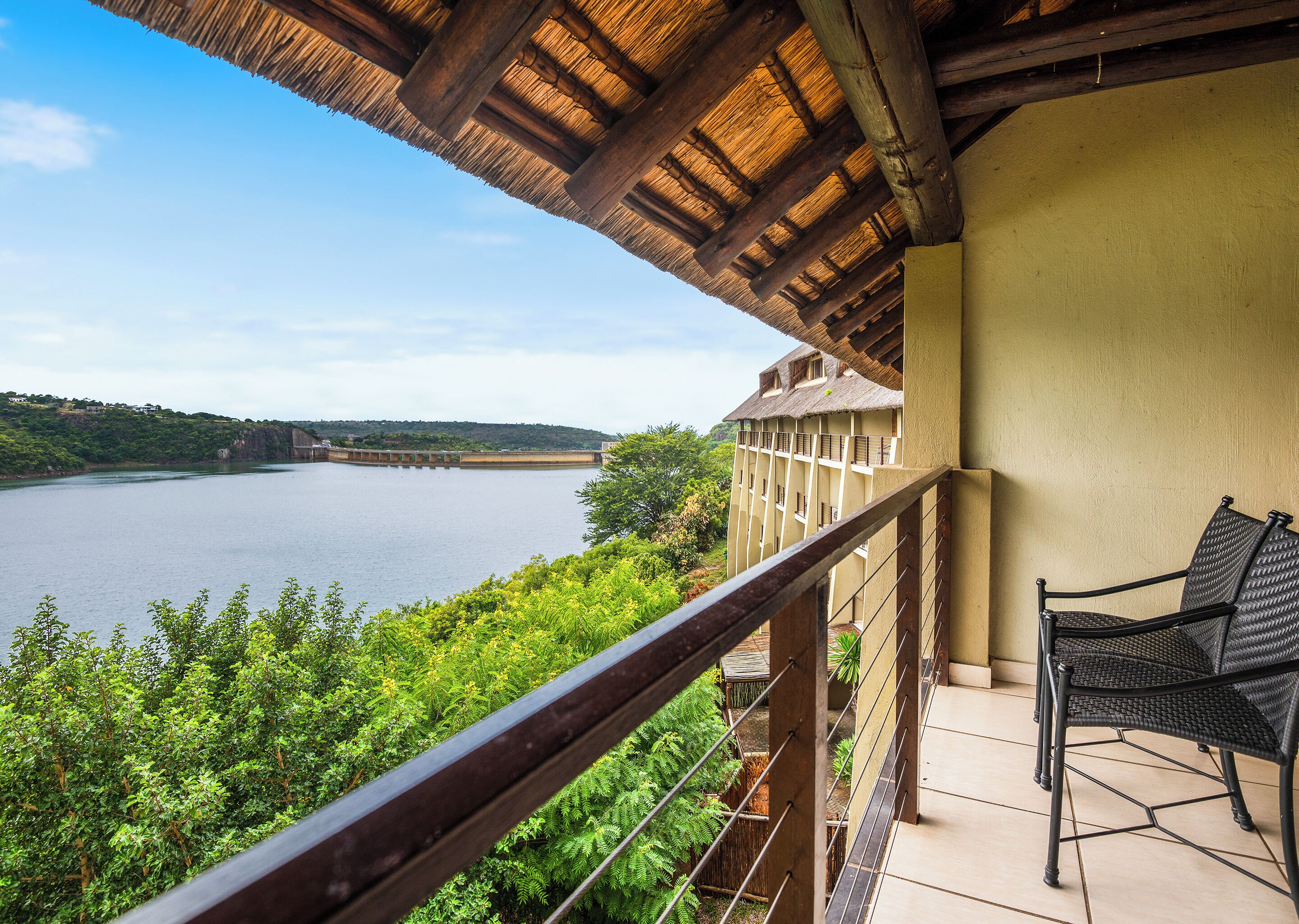 Un balcón con vistas a un lago, rodeado de vegetación y un edificio con tejado de paja.