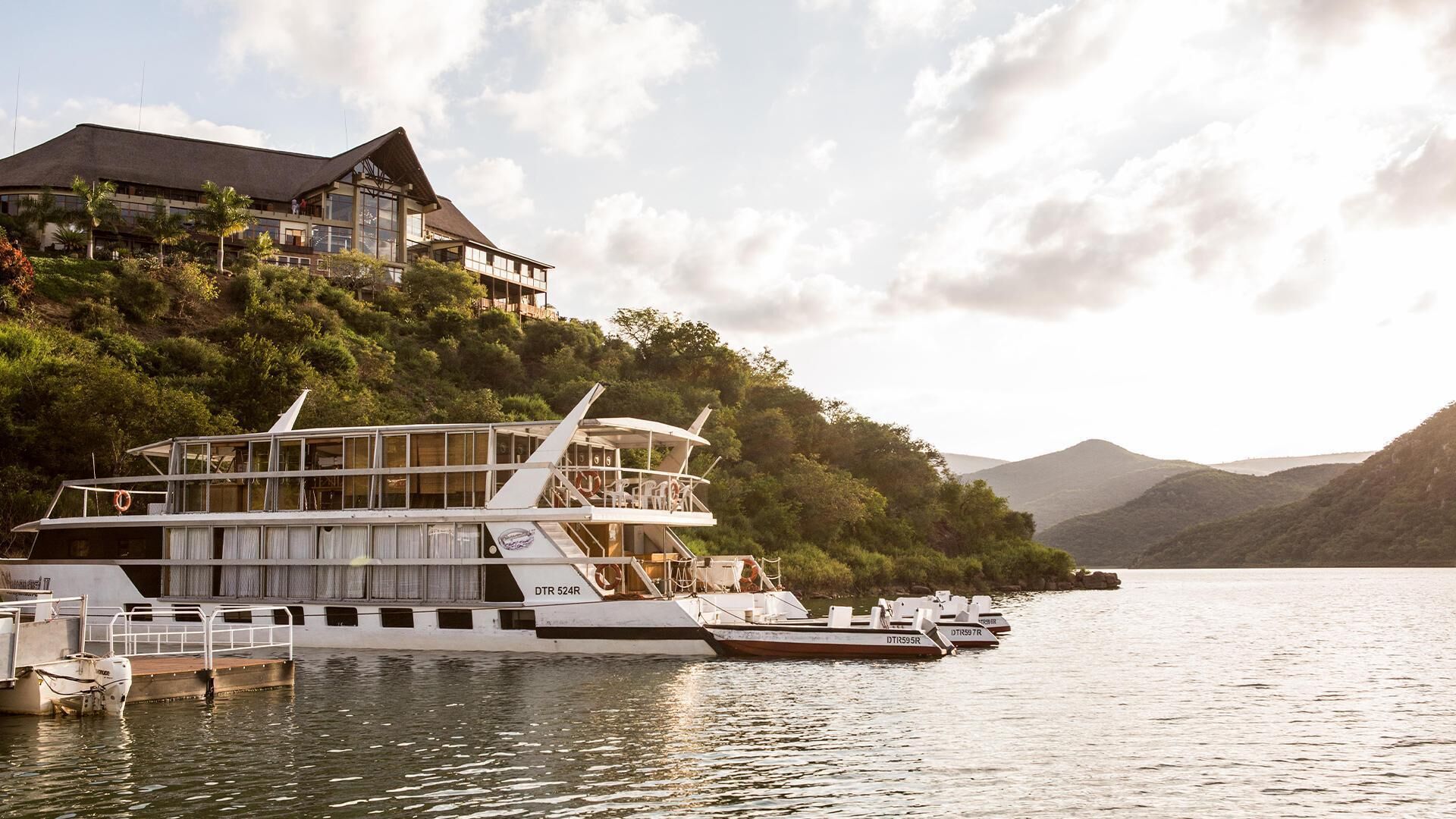 Ein großes, mehrgeschossiges Boot liegt an einem Steg, im Hintergrund erhebt sich ein Haus auf einem Hügel.