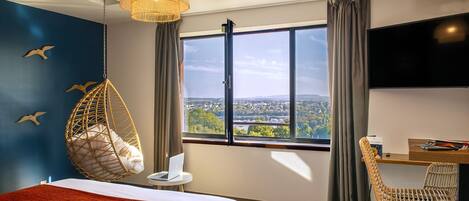 Un dormitorio con cama, escritorio, silla, sillón colgante, televisor y una ventana grande con vista a la ciudad.