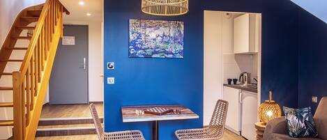 Una sala moderna con una pared azul de acento, una escalera de madera, un comedor con sillas de mimbre y una cocina visible a través de una puerta abierta.