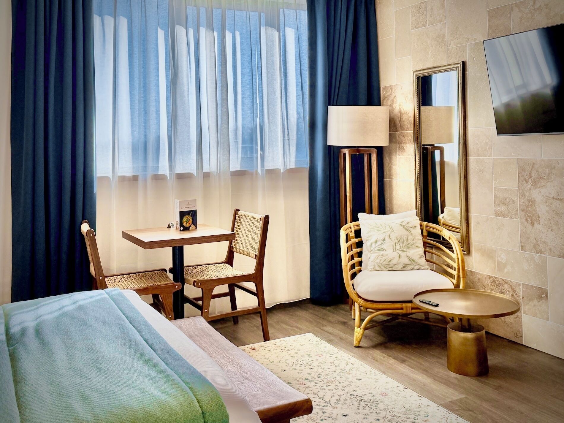 A hotel room with a bed, a desk, two wicker chairs, a small round table, a floor lamp, and a TV mounted on the wall.