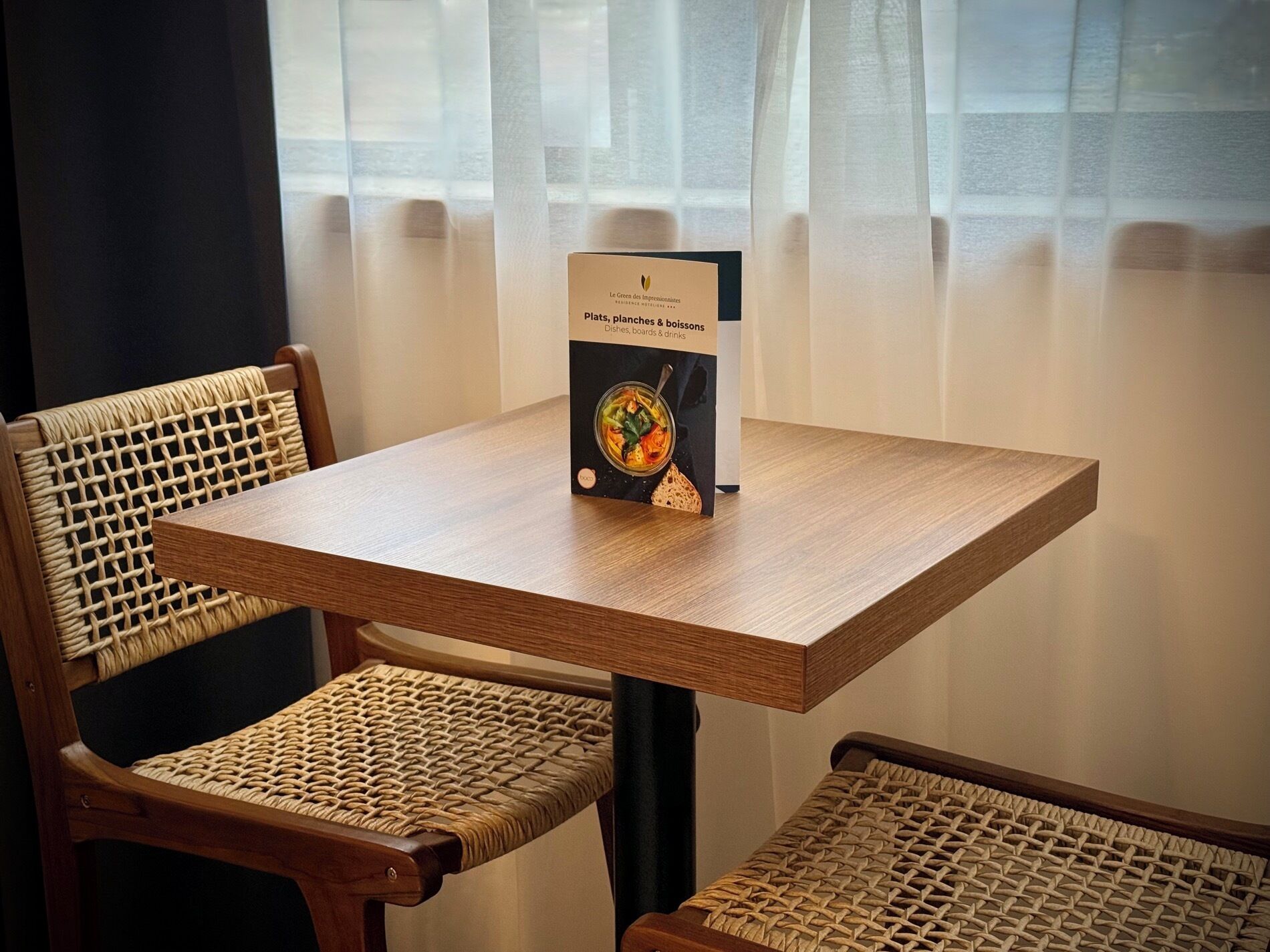 Une table sur laquelle est posé un présentoir à menu, présentant un livre intitulé « Plats, planches et boissons », dans une pièce où se trouvent des chaises en osier et une fenêtre ornée de rideaux légers.