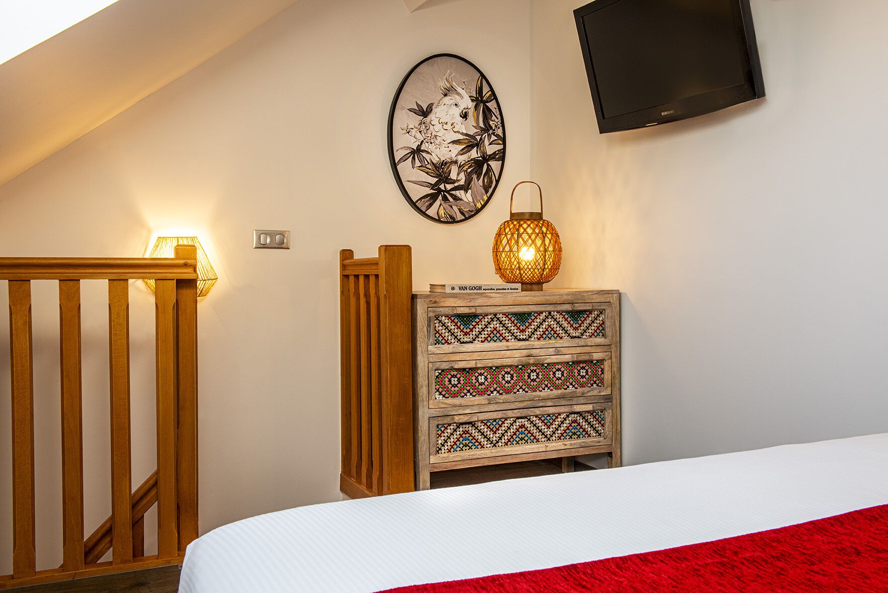 Une chambre à coucher avec un cadre de lit en bois, une couvre-lit blanche, une couverture rouge, un téléviseur fixé au mur, une lampe décorative et une œuvre d’art murale.