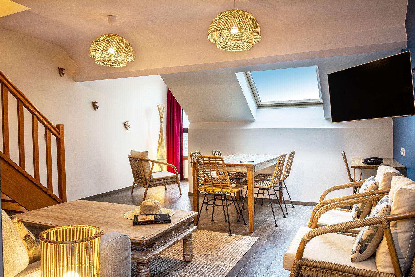 A cozy living room with a dining table, wicker chairs, a sofa, and a TV mounted on the wall.