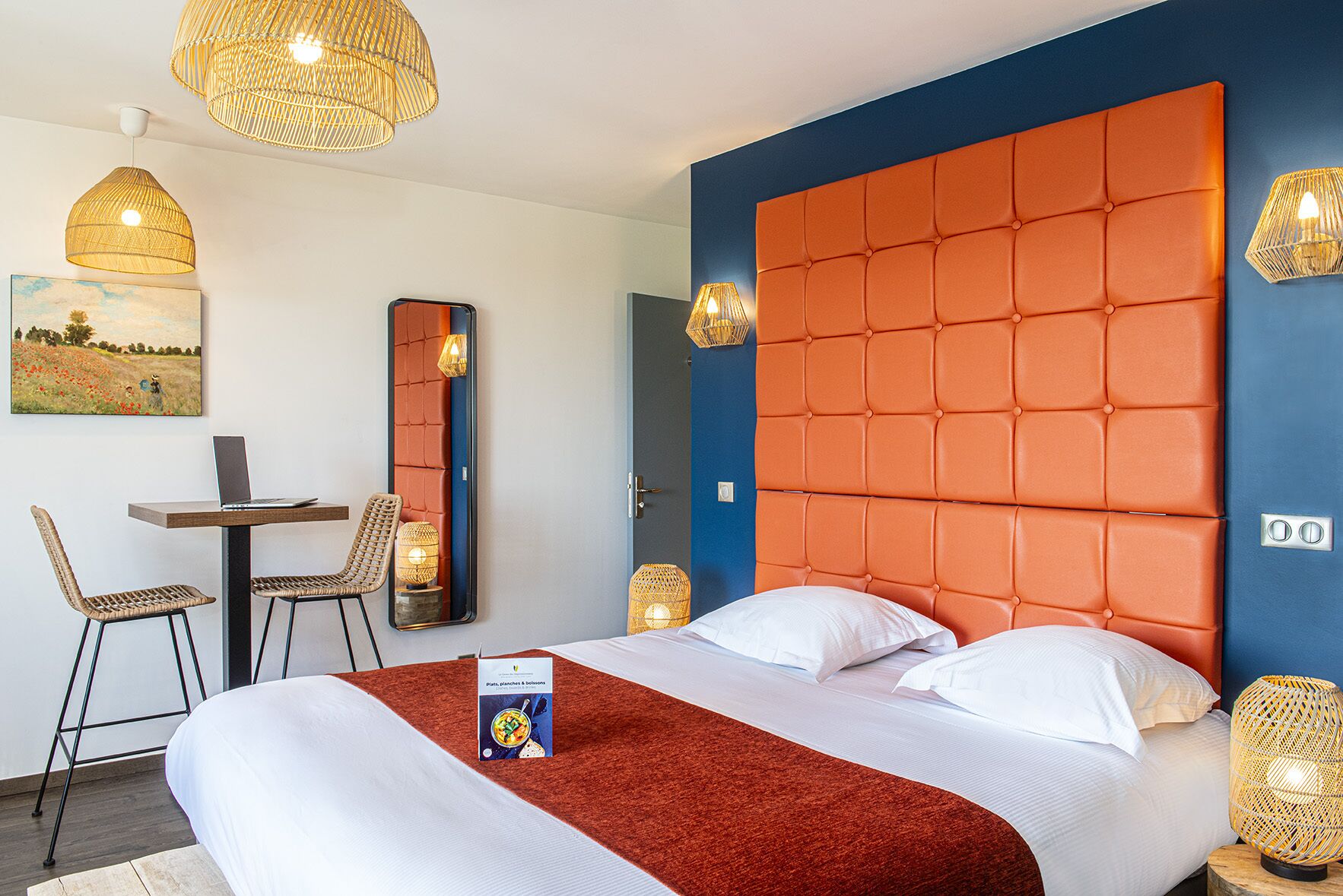 Ein Hotelzimmer mit einem großen Bett, einem Schreibtisch mit einem Laptop, Korbstühlen und einem Bild an der Wand.