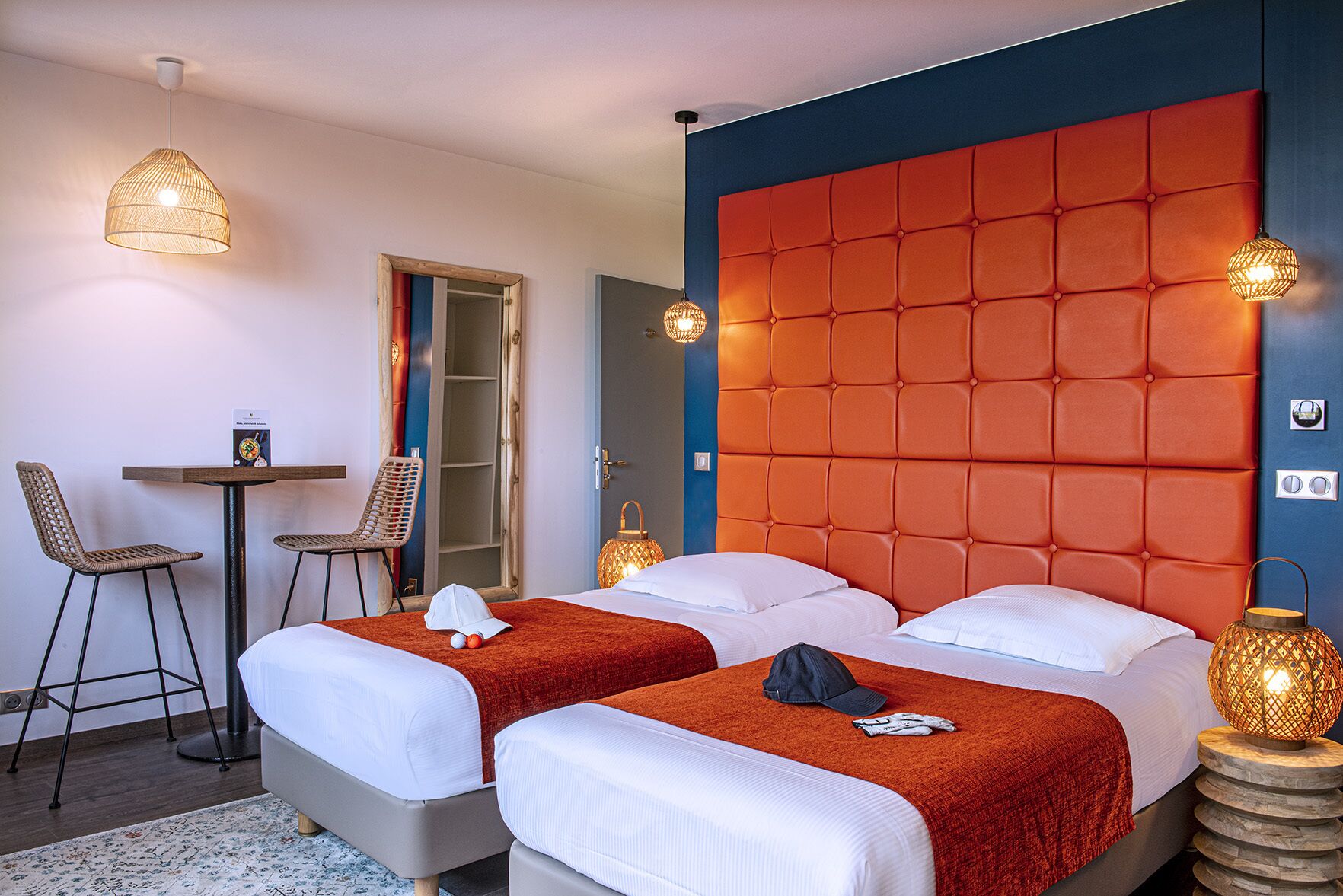 A hotel room with two beds, a small table, two wicker chairs, and a red upholstered headboard.