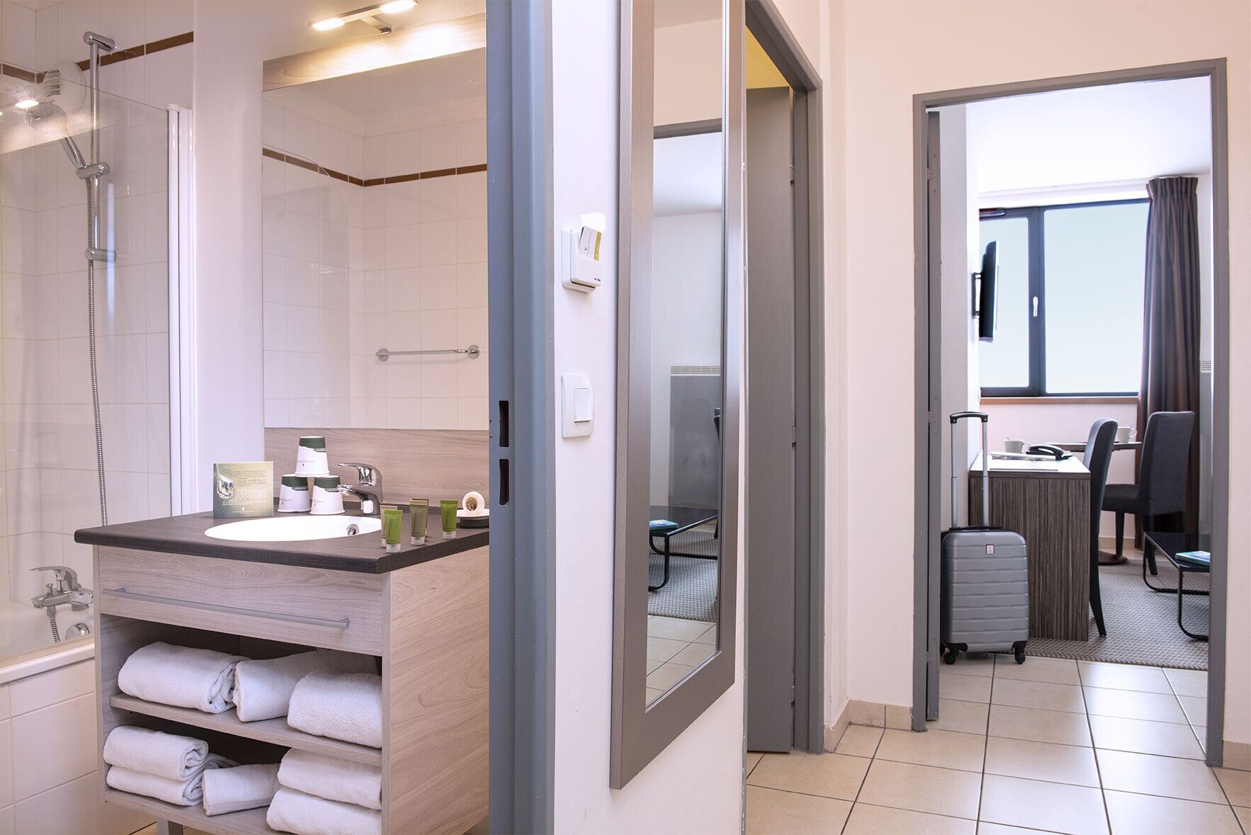 A modern bathroom with a shower, sink, and towel rack. There is a desk and chair in the adjacent room, and a suitcase is visible on the floor.