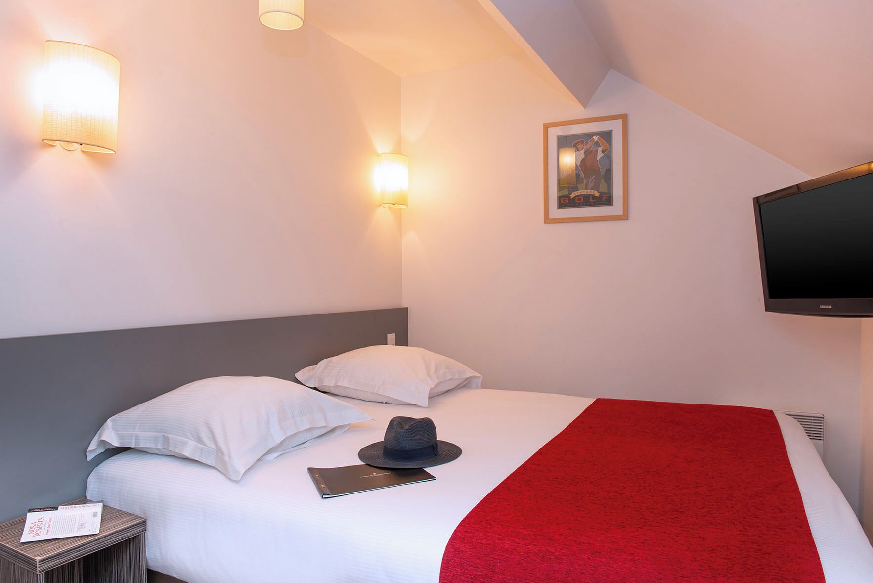 A hotel room with a bed, pillows, a red blanket, a hat, a tablet, and a flat-screen TV.