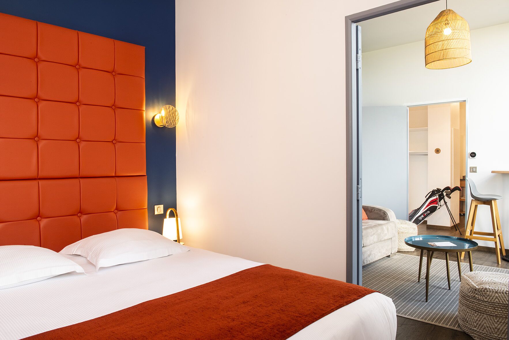 Une chambre d’hôtel avec une tête de lit orange à motifs capitonnés, une couvre-lit blanche, un plaid rouge, une petite table ronde avec une chaise, et un sac de golf en arrière-plan.