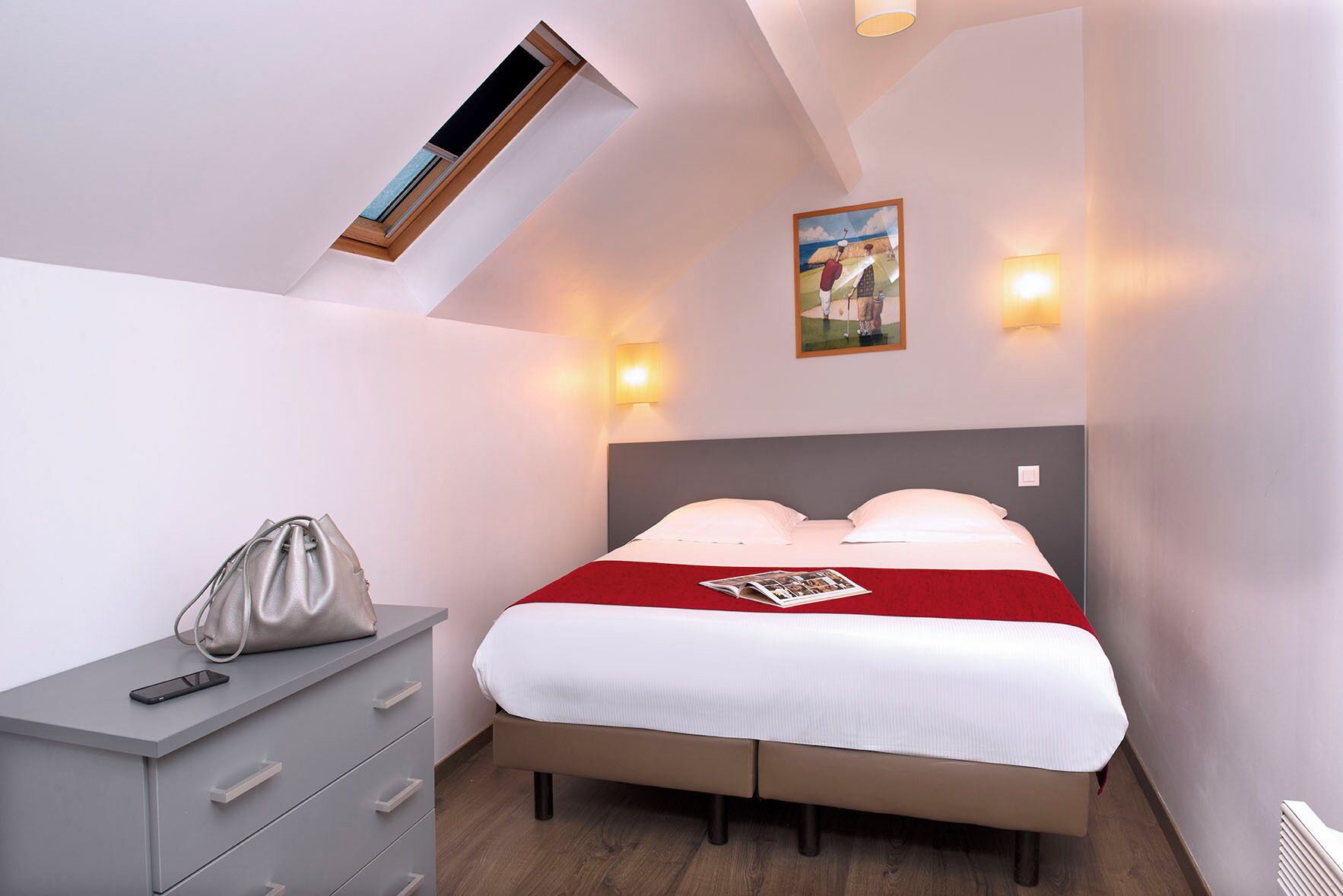 Ein Schlafzimmer mit Bett, Nachttisch, Lampe, Gemälde und Oberlicht.