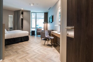 A hotel room with a bed, desk, chair, and a view through a large window. - Sleep & Fly (El Prat de Llobregat)