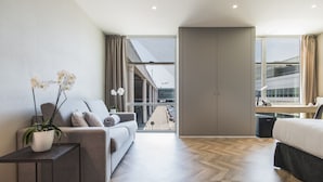 Een moderne hotelkamer met een bank, een nachtkastje met een plant, een bed en uitzicht op de straat en gebouwen.