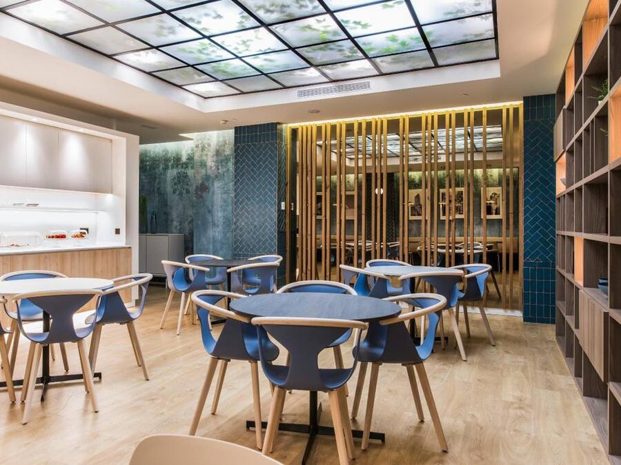 Un espace repas moderne avec des tables rondes et des chaises bleues, une cuisine équipée d’une machine à café et un mur décoratif agrémenté d’éléments verticaux.