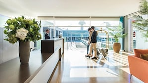 Een moderne hotellobby met een receptiebalie, een grote potplant en uitzicht op het vliegveldterminal.