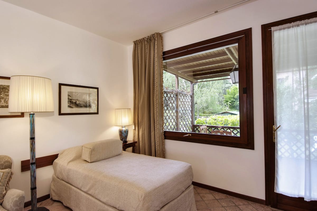 A bedroom with a bed, two bedside lamps, a window with a view of greenery, and a door with a curtain.