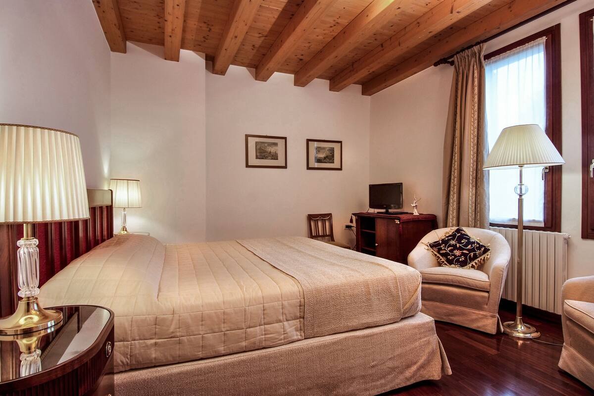 A hotel room with a bed, two bedside lamps, a chair, a desk, and two framed pictures on the wall.