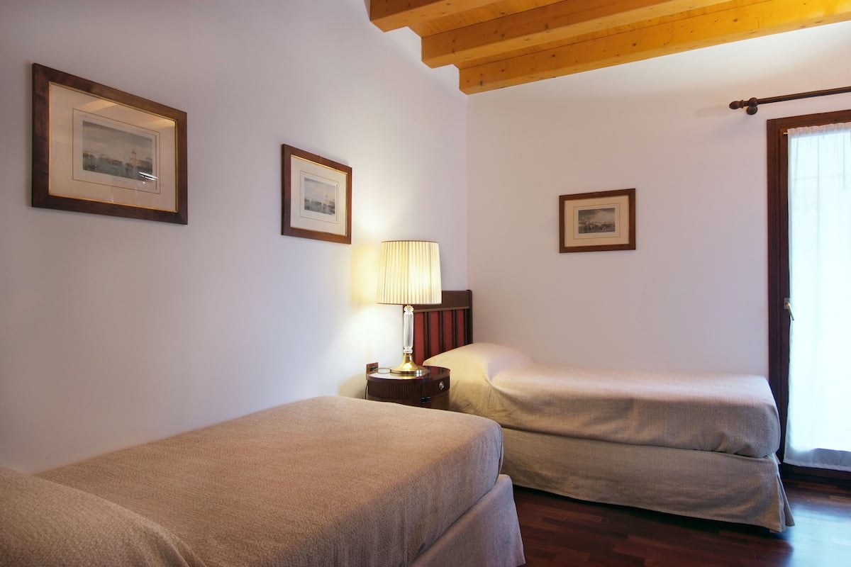A hotel room with two single beds, a nightstand with a lamp, framed pictures on the wall, and a wooden ceiling.