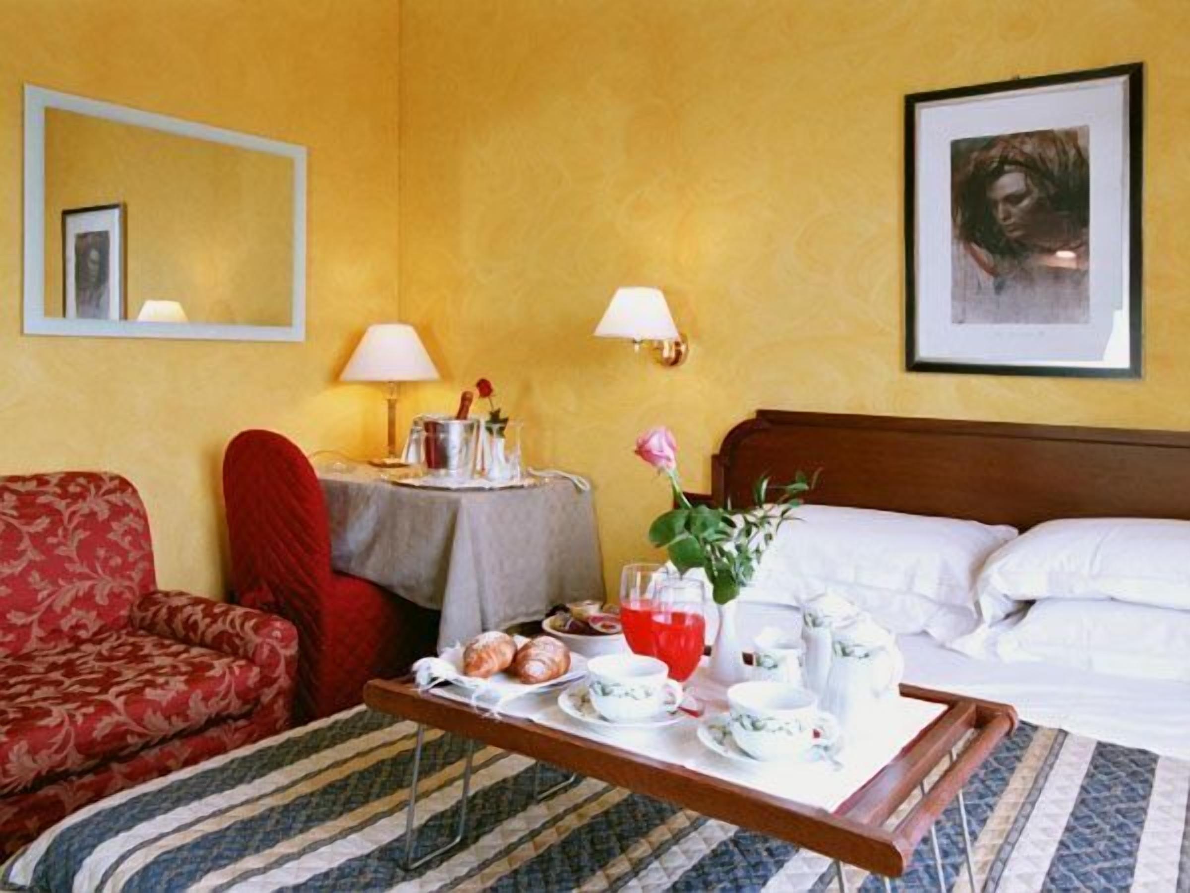 Una habitación de hotel con cama, una mesita de noche con un florero, un sillón rojo, una pequeña mesa con alimentos para el desayuno y un cuadro en la pared.